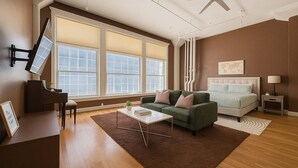 A spacious living room with a piano, a green sofa, a bed, and large windows. - Jazzy Downtown Studio w/ Piano! (Dallas)