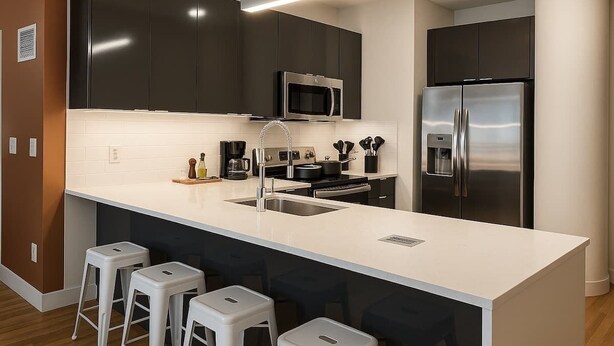 A modern kitchen with dark cabinets, a white countertop, stainless steel appliances, and bar stools. - Jazzy Downtown Studio w/ Piano! (Dallas)