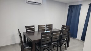 Un comedor con una mesa y sillas de madera oscura, un equipo de aire acondicionado en la pared y un ventanal con cortinas azules.