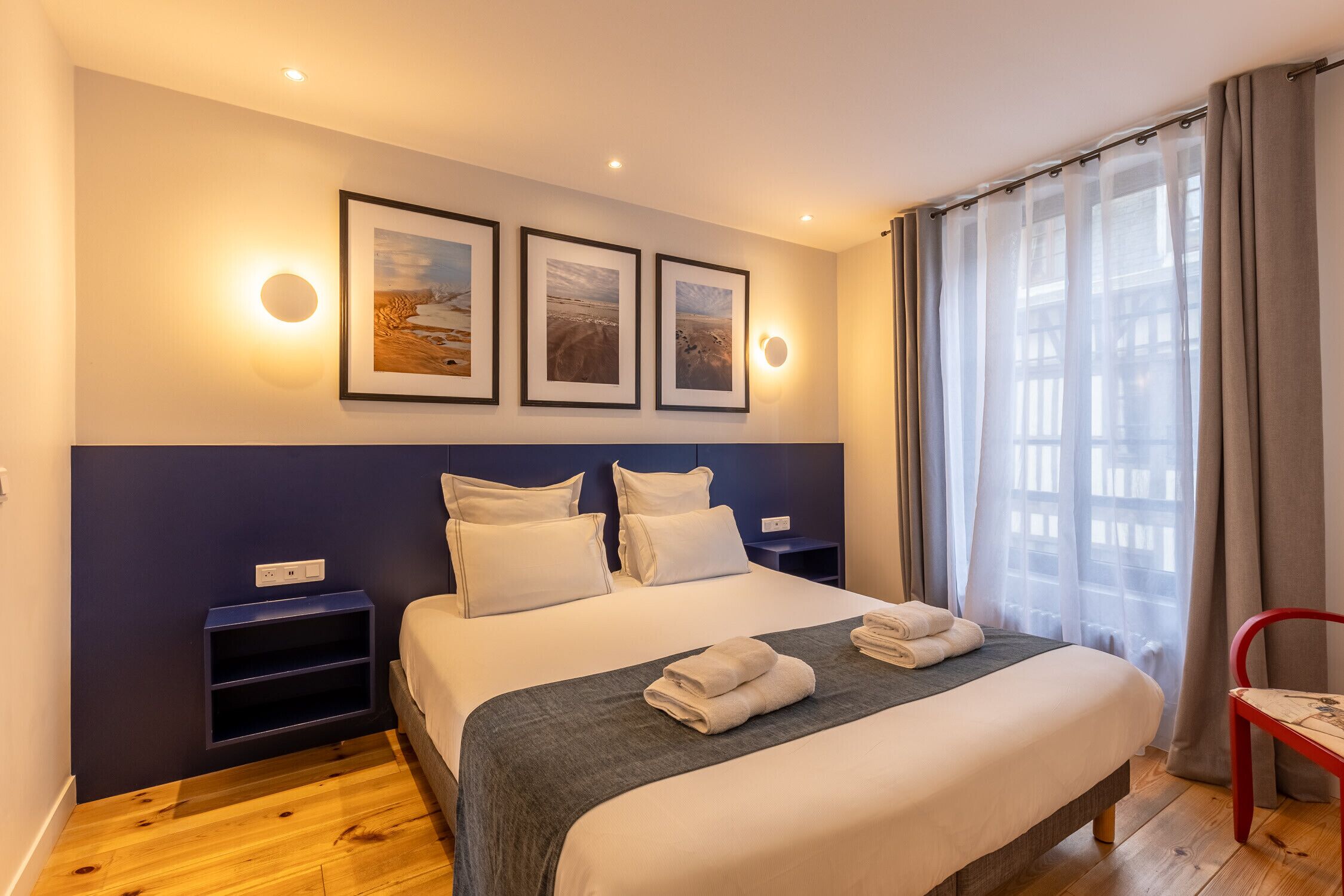 Cama bem arrumada com lençóis brancos e manta cinza, mesa de cabeceira com abajur, e janela com cortinas leves.