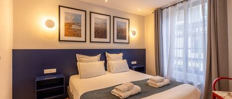 Cama bem arrumada com lençóis brancos e manta cinza, mesa de cabeceira com abajur, e janela com cortinas leves.