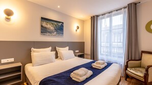 A hotel room with a large bed, a blue throw blanket, two folded towels, a wooden chair, a wall-mounted light, and a painting of a wave. - "Cap sur Honfleur", House in the center of Honfleur 12P (Honfleur)