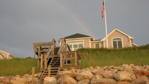 A house with a flag on a pole, a wooden staircase, and a grassy hill. - ''Gulls Way'' - Beachfront and Oceanfront Custom Home on Private Beach (Dennis)