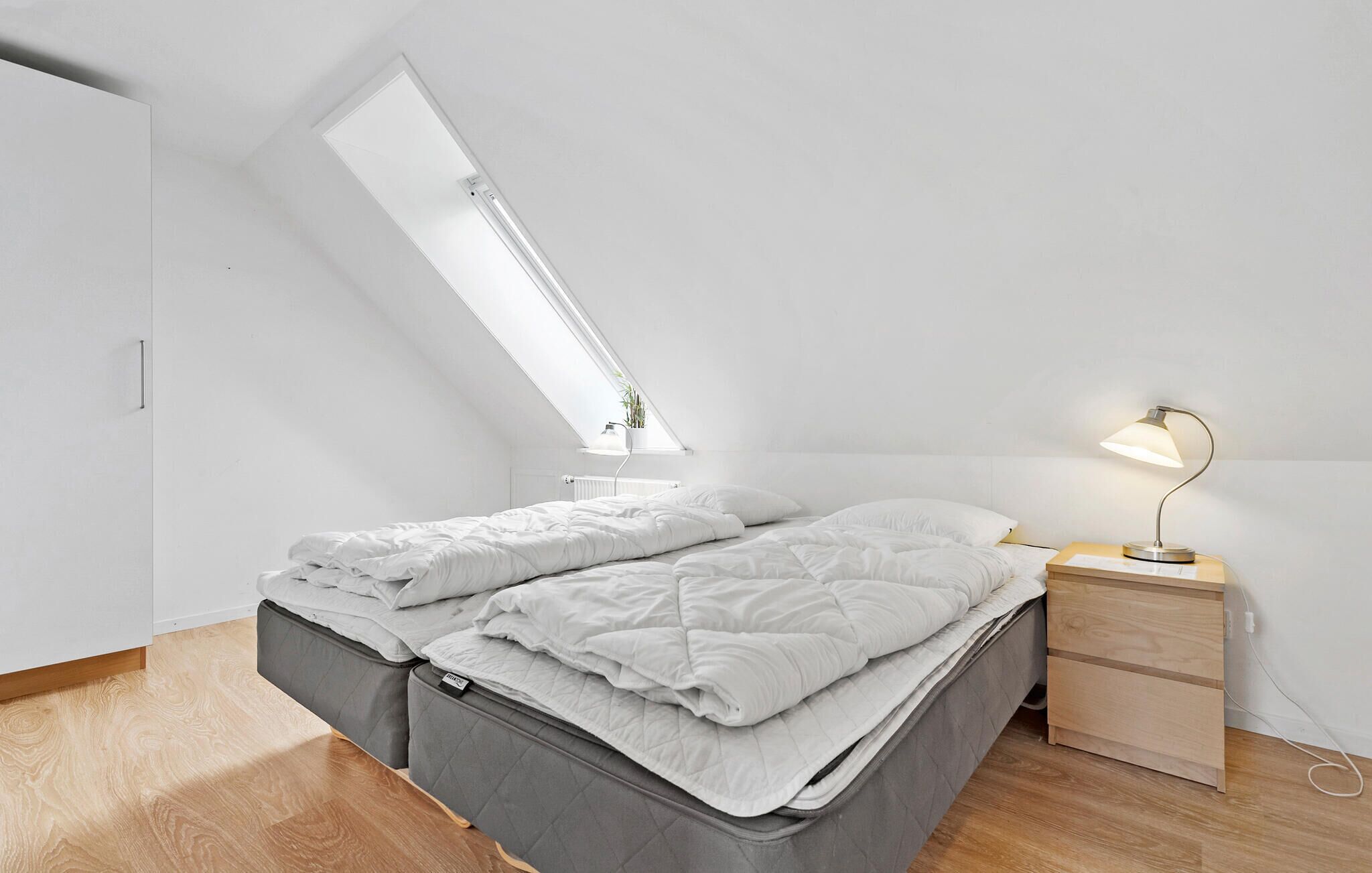 A modern, minimalist bedroom with a sloped ceiling and skylight, a bed with grey bedding, a bedside table with a lamp, and a small plant on the windowsill.