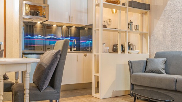 Une cuisine moderne avec des meubles blancs, un four à micro-ondes encastré et une machine à café. Le salon comprend un canapé gris et une petite table avec une chaise.