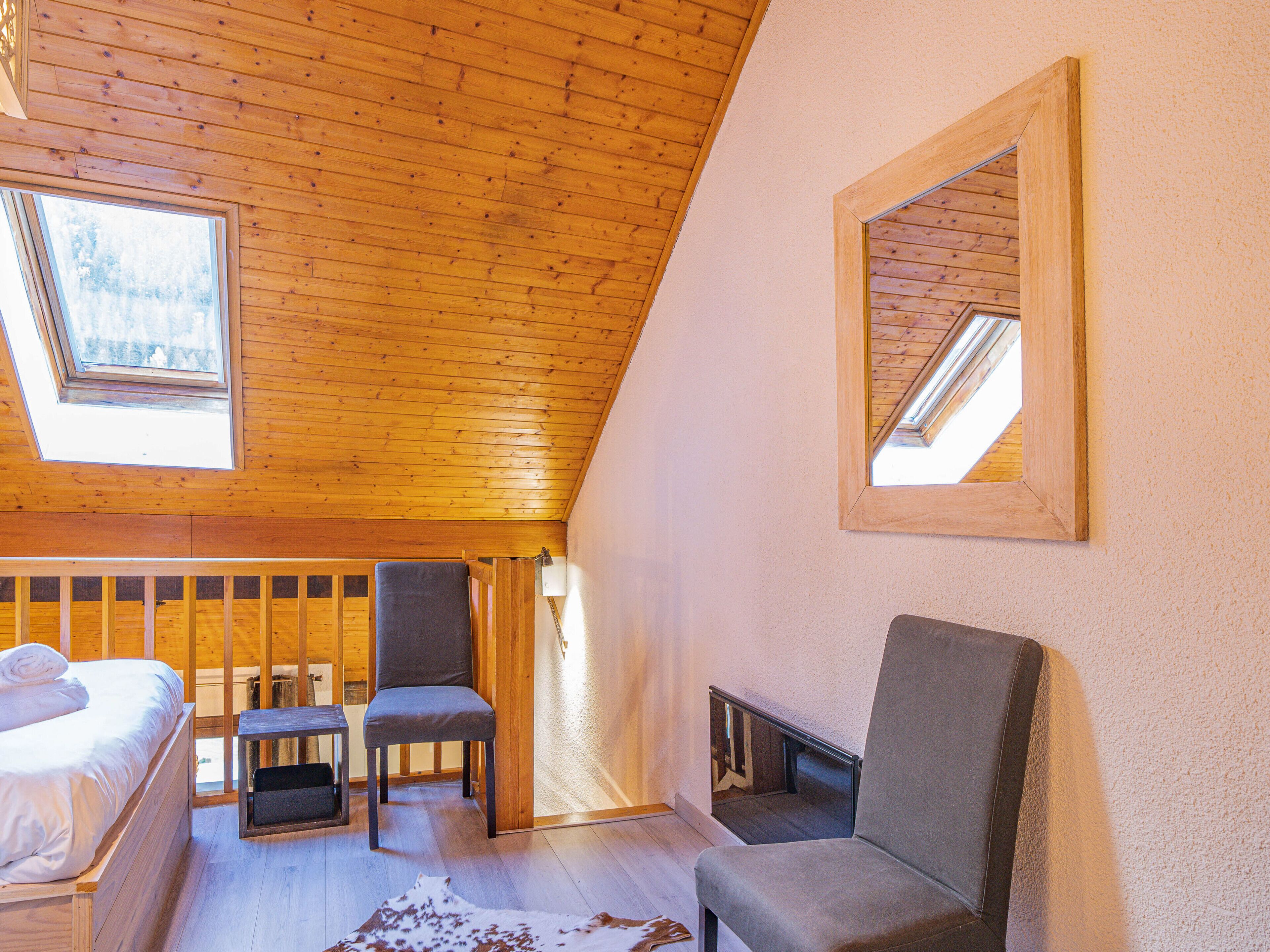 Une pièce chaleureuse avec un plafond en pente, des poutres en bois, un lit, deux chaises, une petite table et une cheminée.