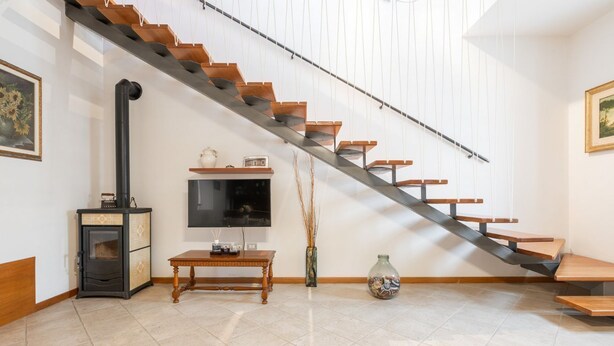 Een moderne woonkamer met een houten trap, een open haard, een flatscreen televisie en een vaas met gedroogde takken.