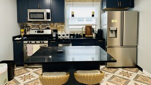 Une cuisine moderne avec des meubles de couleur sombre, des appareils en acier inoxydable, un îlot central doté de deux chaises en osier et un carrelage au motif original.