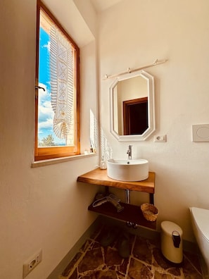 Ein Badezimmer mit einem Holzkosmetikschrank, einem runden Waschbecken, einem Spiegel und einem Fenster mit Blick auf den Himmel.