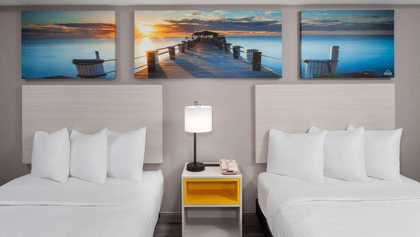 A hotel room with two beds, a nightstand with a lamp, and three paintings of a pier and sunset on the wall.