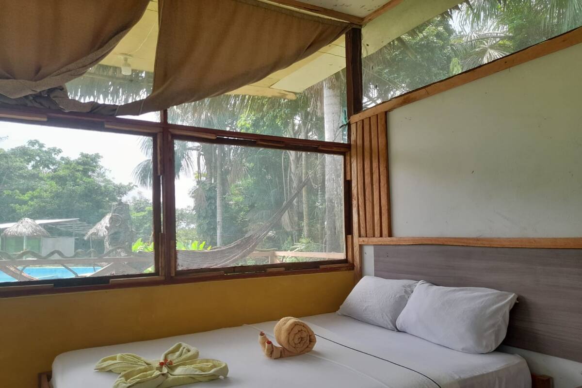 Una cama con colchón blanco, cabecera de madera y vistas a una piscina y árboles a través de un ventanal grande.