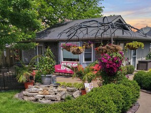 A house with a stone patio, a red bench, hanging flower baskets, and a garden with various plants. - The Cunningham Cottage | King Bed & Garden (Springfield)
