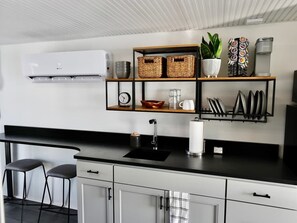 A modern kitchen with a black countertop, white cabinets, a sink, two bar stools, a wall-mounted shelf with baskets and kitchenware, and a wall-mounted air conditioner.