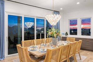 A dining area with a large table set for six, surrounded by wicker chairs, a decorative chandelier, and a vase with flowers.