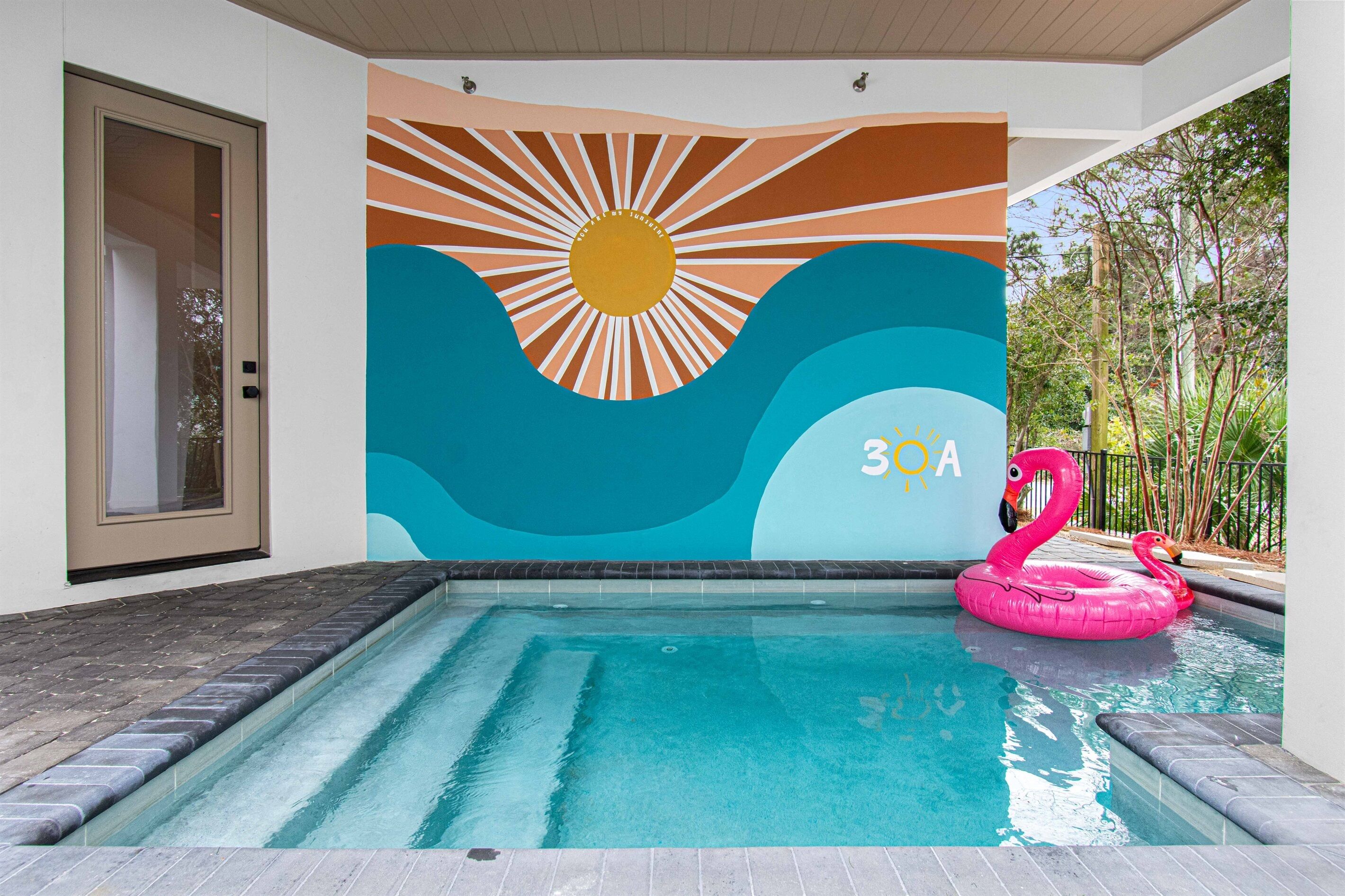 Piscina com um mural de sol e ondas, um boi de flamingo rosa e uma porta à esquerda.