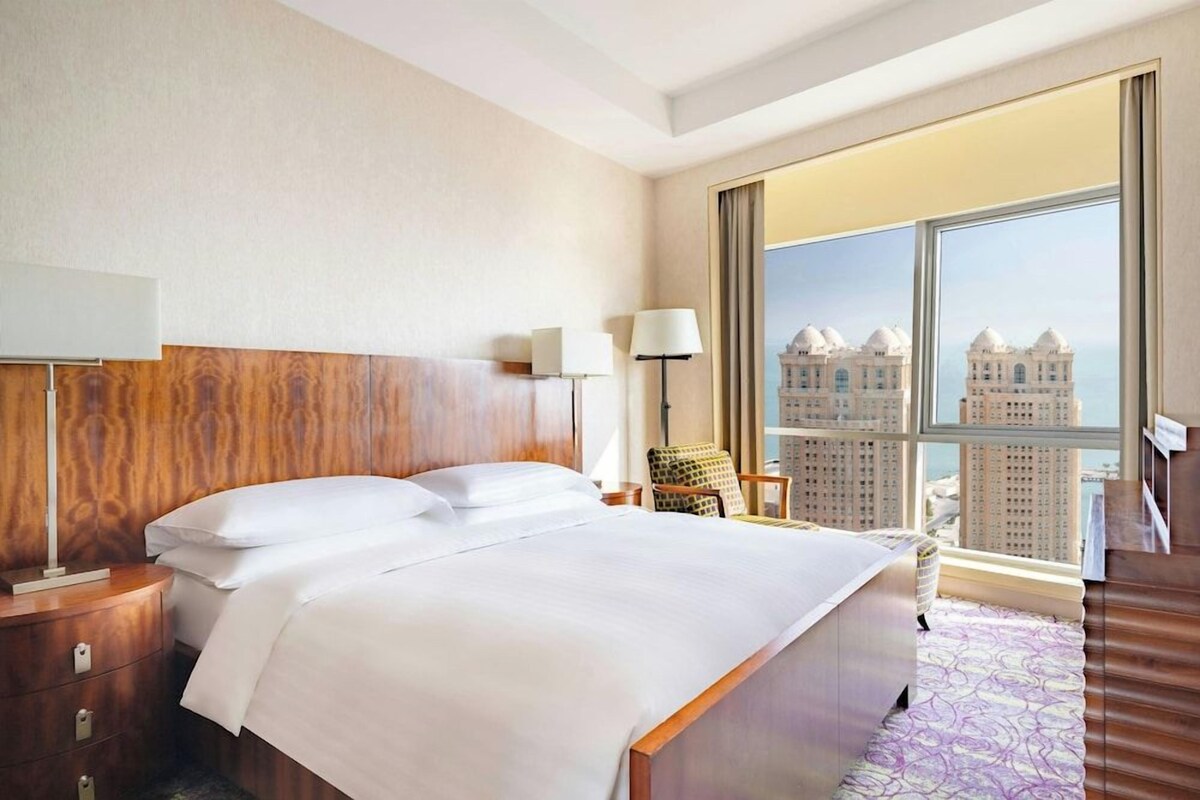 A hotel room with a large bed, a nightstand, a lamp, a chair, and a view of tall buildings through a large window.
