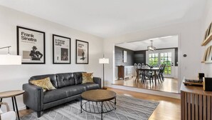 A modern living room with a grey leather sofa, a round coffee table, and a patterned rug. There are framed posters on the wall, a dining area with a wooden table and chairs, and a kitchen visible through an open doorway.