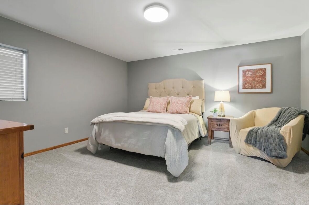 Een slaapkamer met een bed, nachtkastje, stoel en een ingelijst kunstwerk aan de muur.