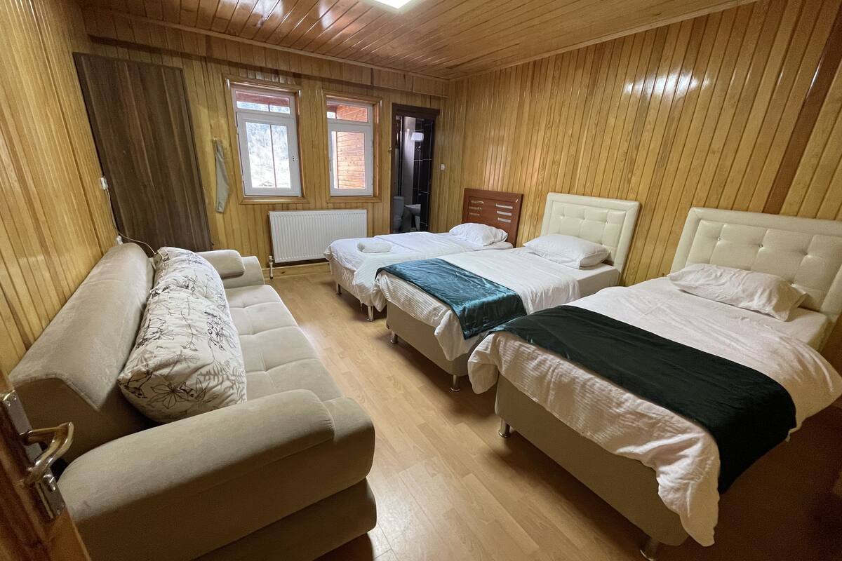 Ein Zimmer mit Holzwänden, zwei Betten mit weißen Leinentüchern, einem beigen Sofa und einem Fenster mit Jalousien.