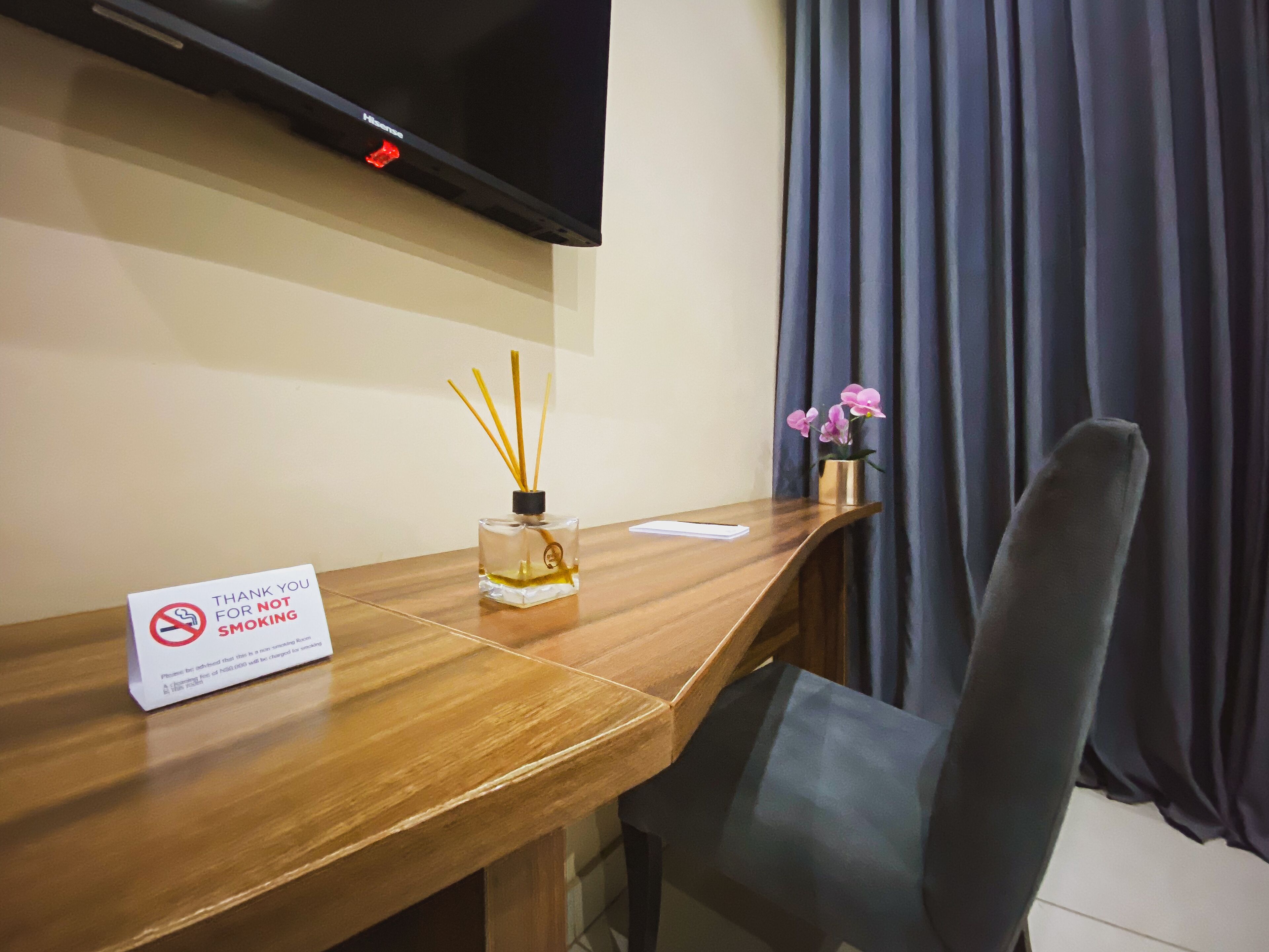 A hotel room with a wooden desk, a television mounted on the wall, a sign prohibiting smoking, a vase with pink flowers, and a chair.