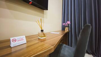 A hotel room with a wooden desk, a television mounted on the wall, a sign prohibiting smoking, a vase with pink flowers, and a chair.