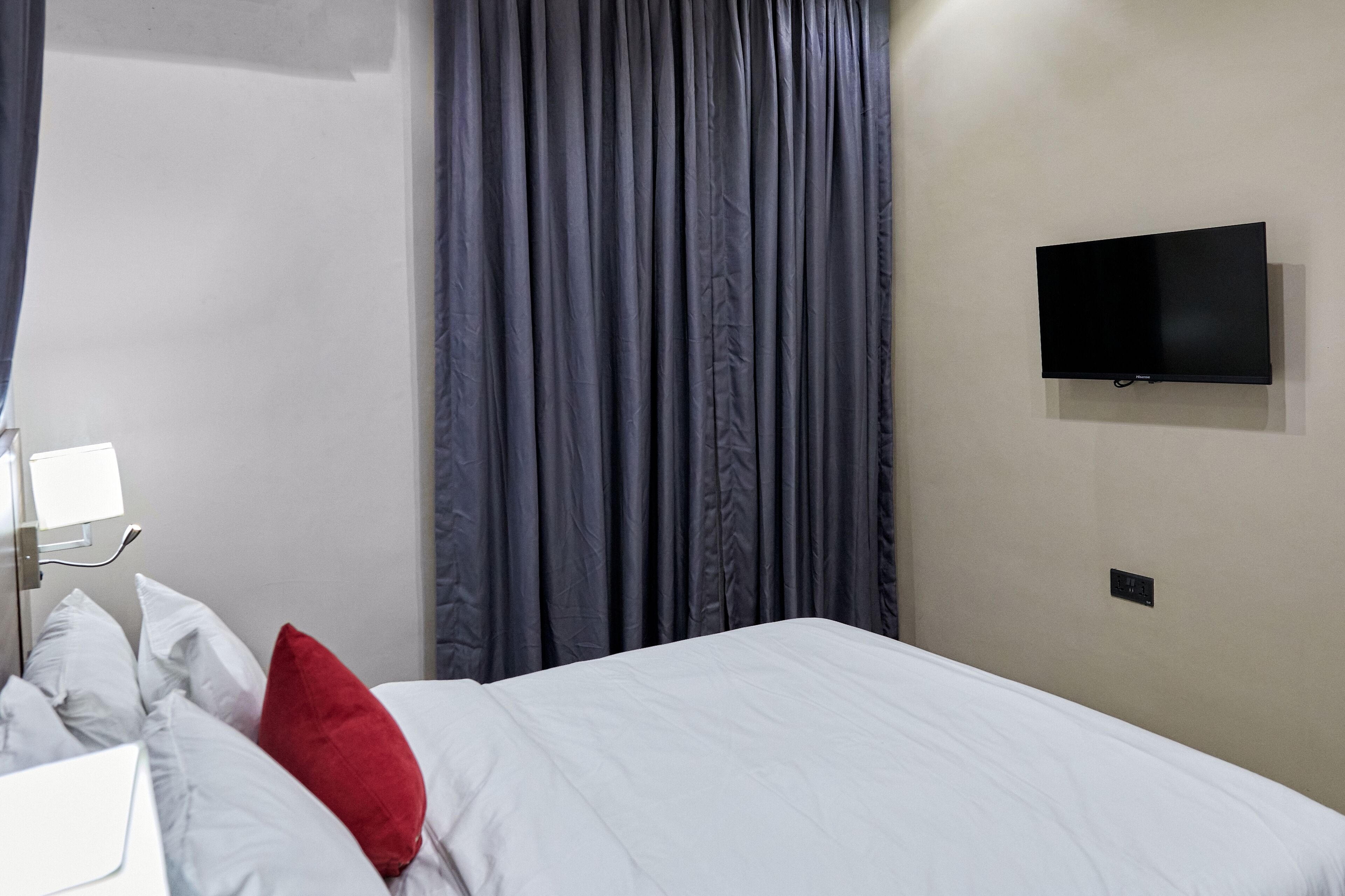 Habitación de hotel con cama, ropa de cama blanca, almohada roja, televisor de pantalla plana montado en la pared y una ventana con cortinas.