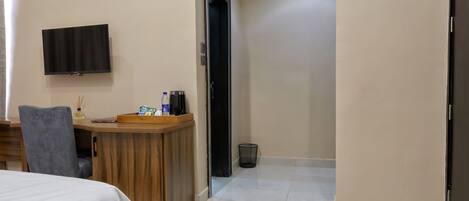 A hotel room with a bed, a desk with a chair, a television mounted on the wall, and a door leading to another room.