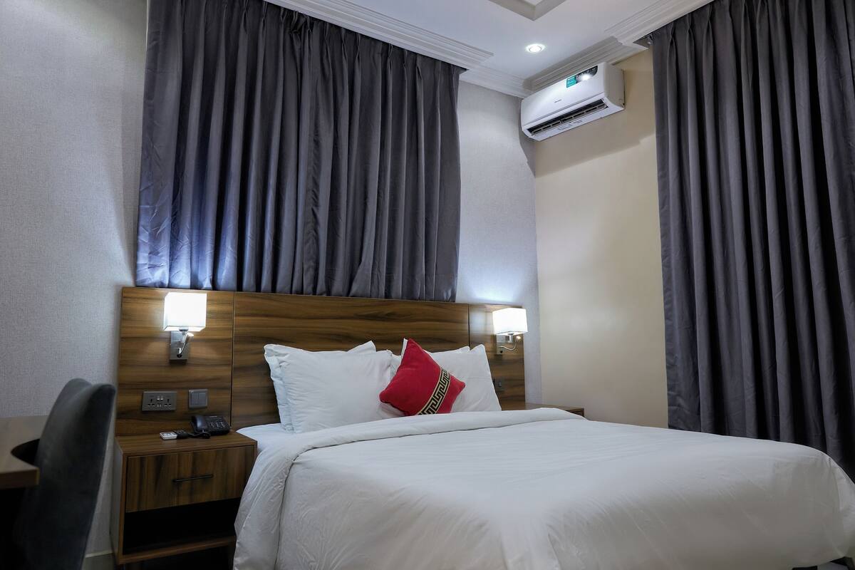 Une chambre d’hôtel avec une tête de lit en bois, du linge de lit blanc, un coussin décoratif rouge, une lampe de chevet et un appareil de climatisation.