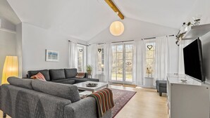 Un salon moderne avec un canapé gris, une table basse et un téléviseur à écran plat. De grandes fenêtres, ornées de rideaux, offrent une vue sur les arbres à l’extérieur.