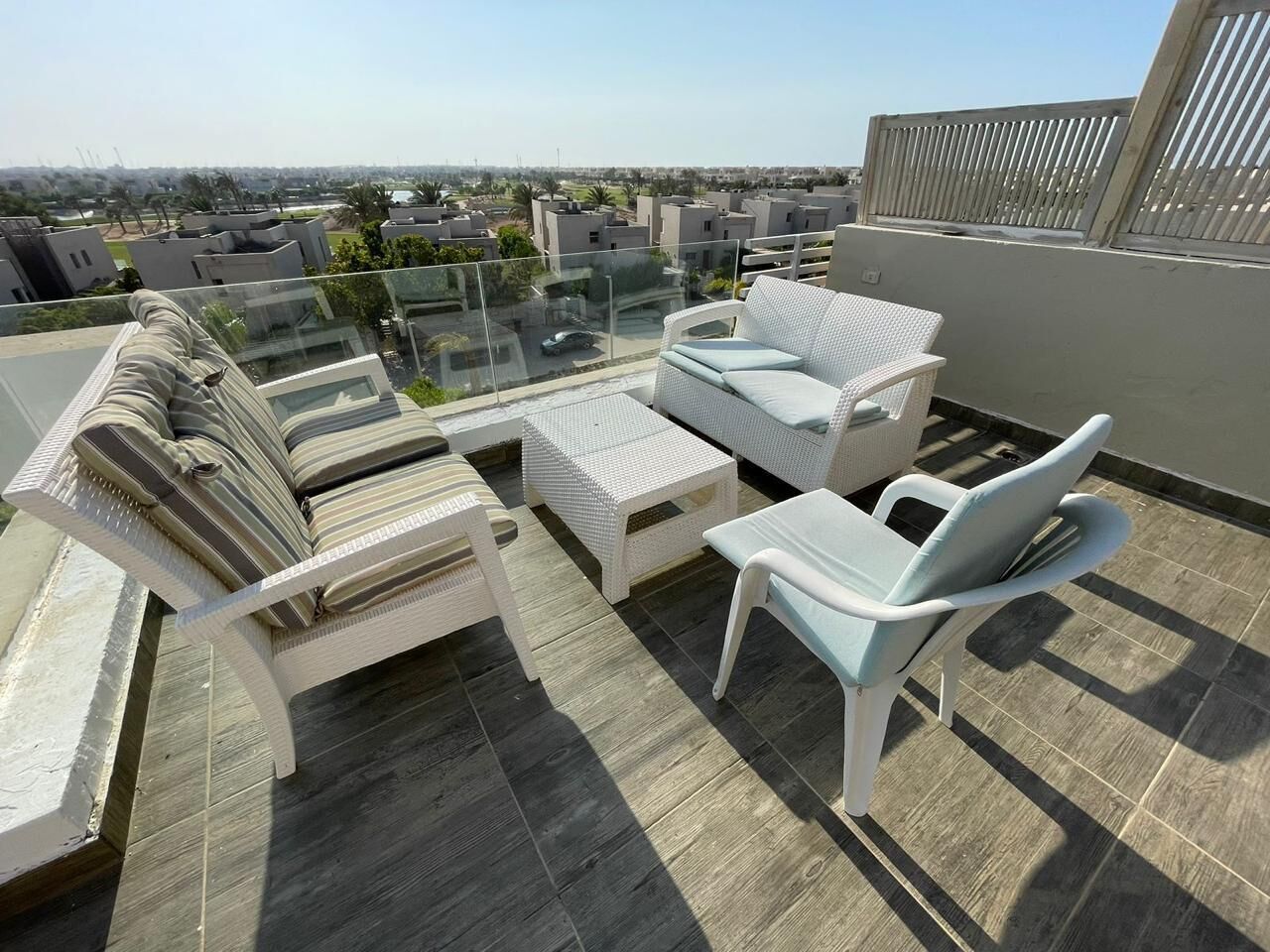 A rooftop terrace with a glass balustrade, white wicker furniture, and a view of the cityscape.