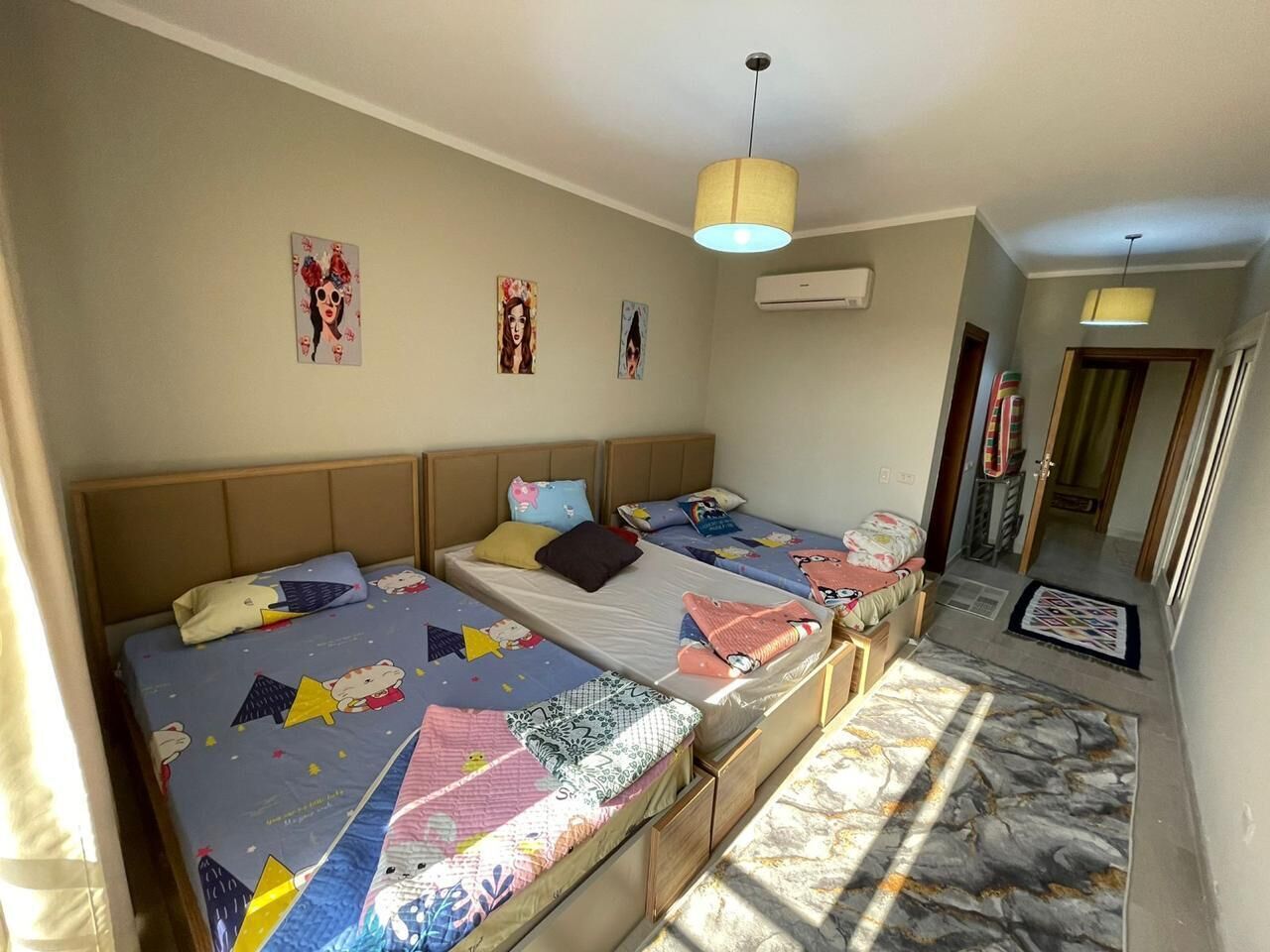 Habitación de hotel con tres camas, un ventanal grande y un pasillo que lleva a una puerta.