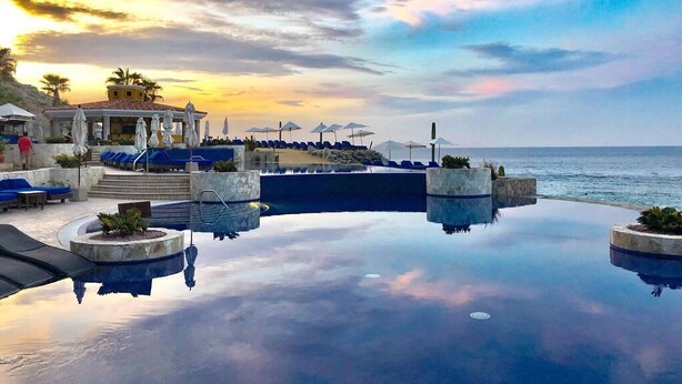 Ein luxuriöser Infinity-Pool mit klarem Wasser, das den Himmel spiegelt, umgeben von blauen Liegestühlen und Sonnenschirmen, mit Blick auf den Ozean und ein Gebäude mit einem Ziegeldach im Hintergrund.