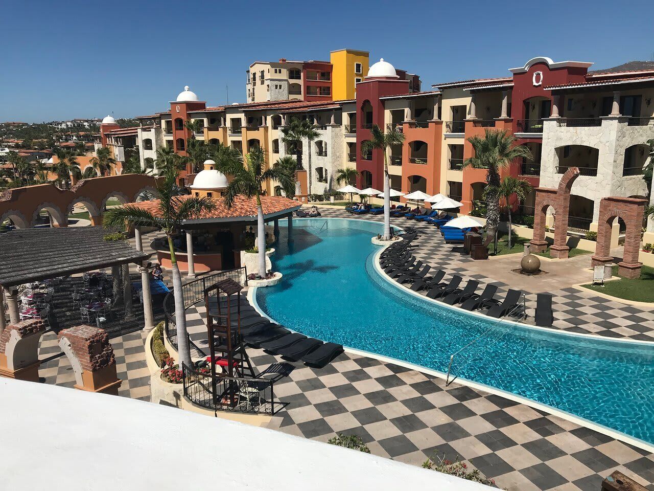 Een resort met een zwembad in een boogvorm, ligbedden en een geruite vloer.