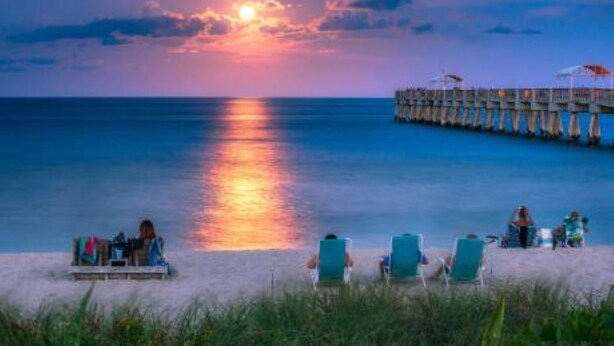 Una playa con un muelle que se adentra en el agua, un atardecer de fondo y varias personas relajándose en la arena.