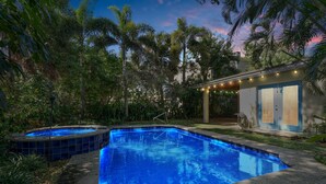Una piscina con borde de baldosas, rodeada de vegetación exuberante y una casa con luces de cadena.