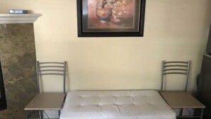Ein kleines Zimmer mit einem Einzelbett, zwei Stühlen, einem gerahmten Bild an der Wand und einer Steinwand.