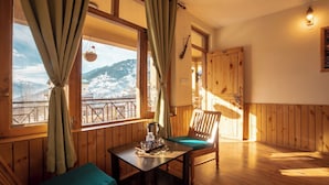 Une pièce chaleureuse aux murs et au plancher en bois, avec une table, une bouilloire, une chaise et une vue sur des montagnes enneigées, visible à travers une grande fenêtre ornée de rideaux.