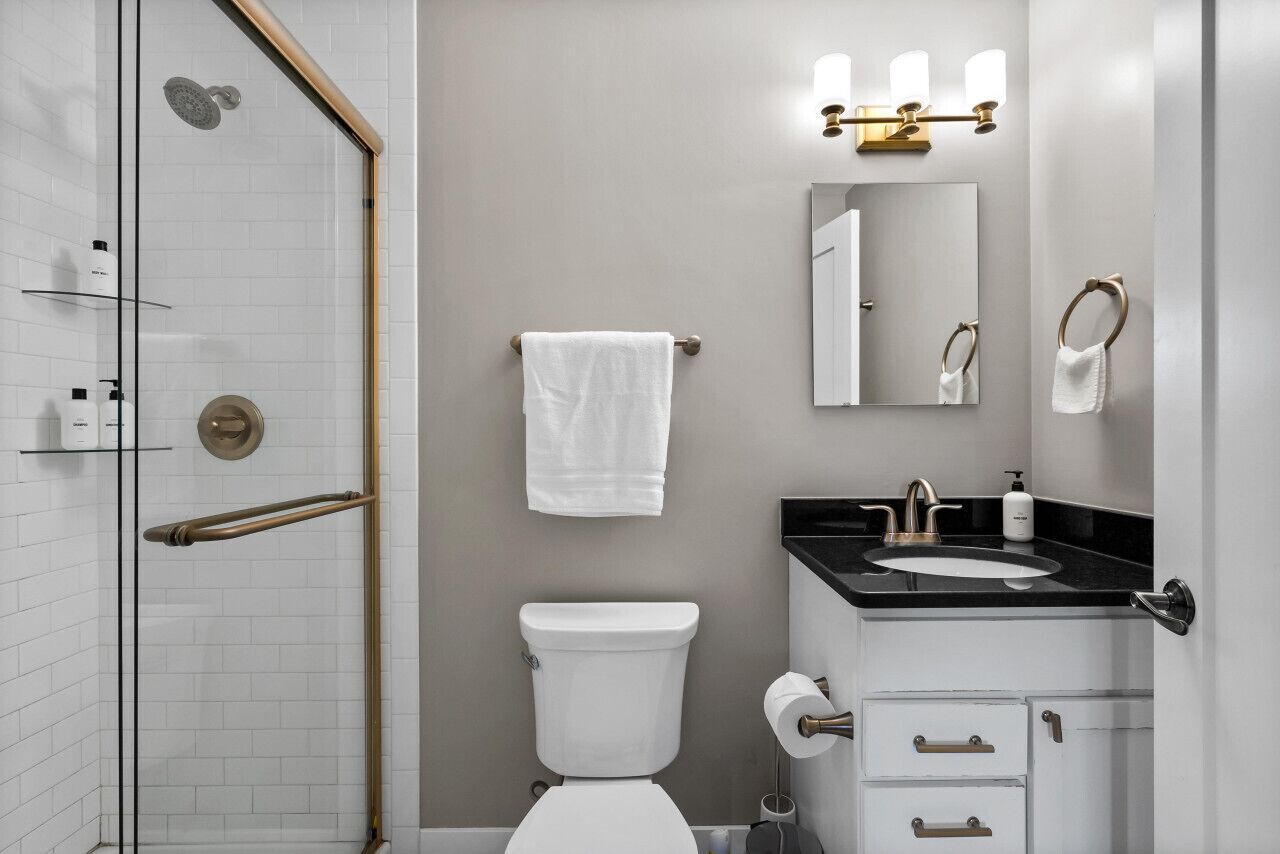 Un baño con cabina de ducha de vidrio, inodoro, lavabo con encimera negra y espejo con luces empotradas.
