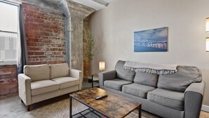 A living room with a brick wall, two sofas, a coffee table, and a cityscape painting. - The Nelson Lofts - Unit 4 (2 BR) (Davenport)