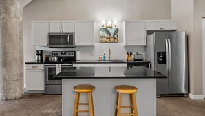 A modern kitchen with white cabinets, stainless steel appliances, a central island with stools, and a decorative painting above the sink. - The Nelson Lofts - Unit 4 (2 BR) (Davenport)