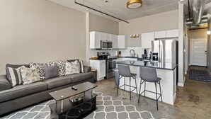 A compact living space with a kitchenette, a sofa with patterned cushions, a coffee table, and a bar area with stools. - The Nelson Lofts - Unit 6 (1 BR) (Davenport)
