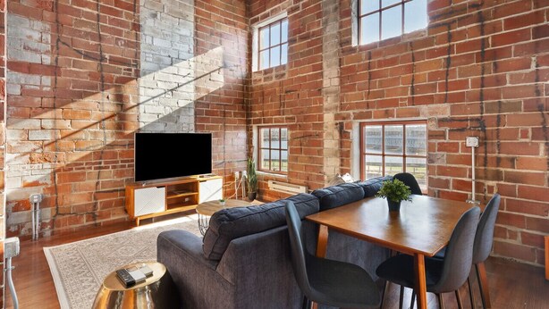 A spacious living area with a brick wall, a large window, a flat-screen TV, a sofa, a dining table, and a staircase. - The Nelson Lofts Penthouse (2BR) (Davenport)