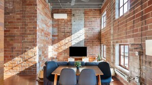 A room with a brick wall, a wooden table with chairs, a sofa, and a TV. - The Nelson Lofts Penthouse (2BR) (Davenport)
