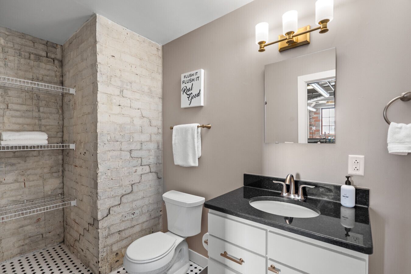 Un baño con pared decorativa de ladrillo, inodoro blanco, lavabo negro con grifo dorado, espejo y toallero.