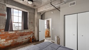 Une pièce aux murs en briques apparentes, une grande fenêtre avec des rideaux, un lit avec une couette grise et une armoire blanche.