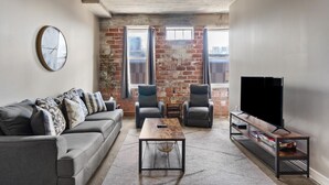 A modern living room with a grey sofa, two armchairs, a coffee table, and a TV on a wooden stand. - The Nelson Lofts - Unit 3 (2 BR) (Davenport)