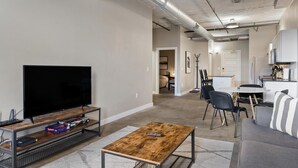A modern living room with a flat-screen TV, a coffee table, and a sofa. There is a dining area with a table and chairs. The room has a neutral color palette and features exposed ductwork on the ceiling. - The Nelson Lofts - Unit 3 (2 BR) (Davenport)