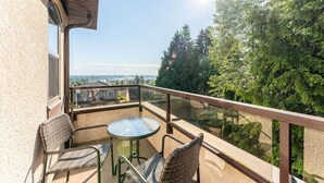 Un balcon doté d’une table en verre et de deux fauteuils en osier, offrant une vue sur le paysage urbain et une forêt dense.