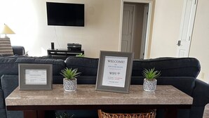 A living room with a dark-colored sofa, a coffee table with a registration certificate, a welcome sign, and two potted plants.