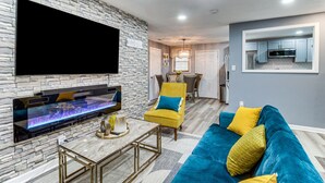 A modern living room with a stone wall, a flat-screen TV, a fireplace, a yellow armchair, a blue sofa, and a kitchen area in the background. - Inviting Savannah Home w/ Yard: 3 Mi to Downtown! (Savannah)